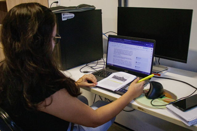 Os servidores poderão cumprir a jornada diária de oito horas, divididas em seis horas para atividades administrativas e duas horas para participação em cursos de capacitação (Foto: Ribamar Neto/UFC Informa) Imagem: Fotografia de uma pessoa sentada diante de uma mesa de trabalho em um escritório. Vista de costas e de perfil, ela utiliza um notebook enquanto consulta um documento impresso aberto ao lado do computador, segurando um marcador de texto amarelo. Na tela do notebook, há um editor de texto com um documento em elaboração. Sobre a mesa também estão um mouse, um monitor desligado, um computador de mesa, um smartphone e um caderno. O ambiente é iluminado por luz artificial e sugere uma atividade de revisão ou produção de documentos.