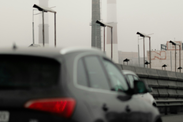 Imagem: cenário urbano industrial sob neblina. Ao fundo, destacam-se três chaminés altas de uma fábrica, parcialmente encobertas pela névoa. Em primeiro plano, aparece um carro escuro visto de trás, desfocado, circulando em uma via elevada com postes de iluminação ao longo da pista. O clima é frio e poluído, com baixa visibilidade.